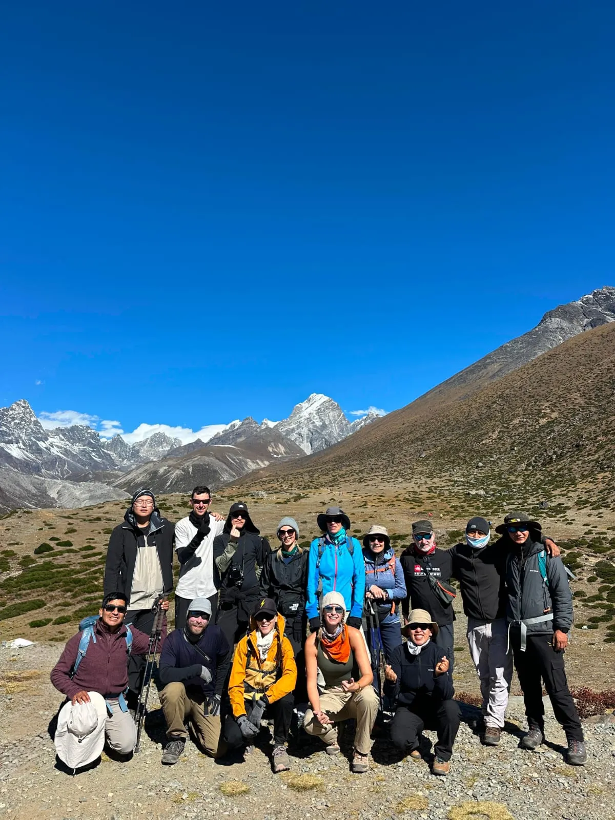A large trekking group posing on a brown hillside below snowy mountains