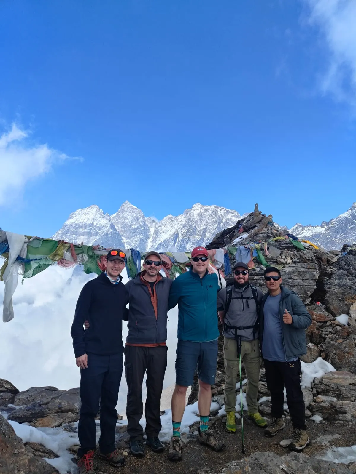 A group of five trekkers under prayer flags, with snow-capped peaks behind