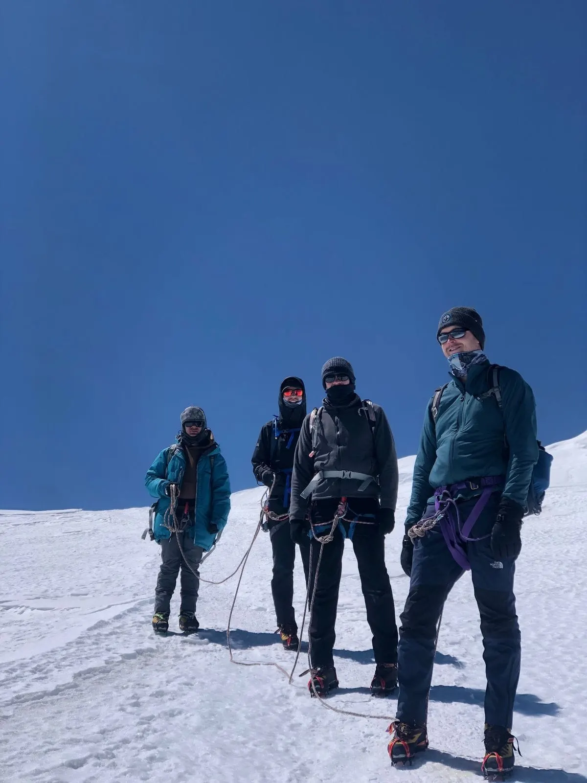 Four roped climbers in harnesses standing on snow under a clear blue sky