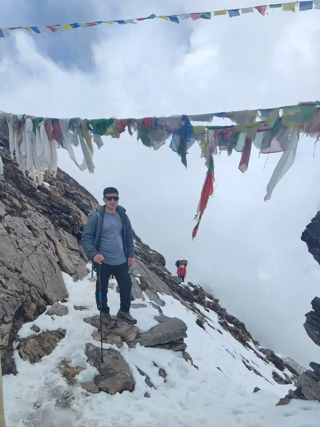A trekker standing on rocks under prayer flags, with cloud filling the valley