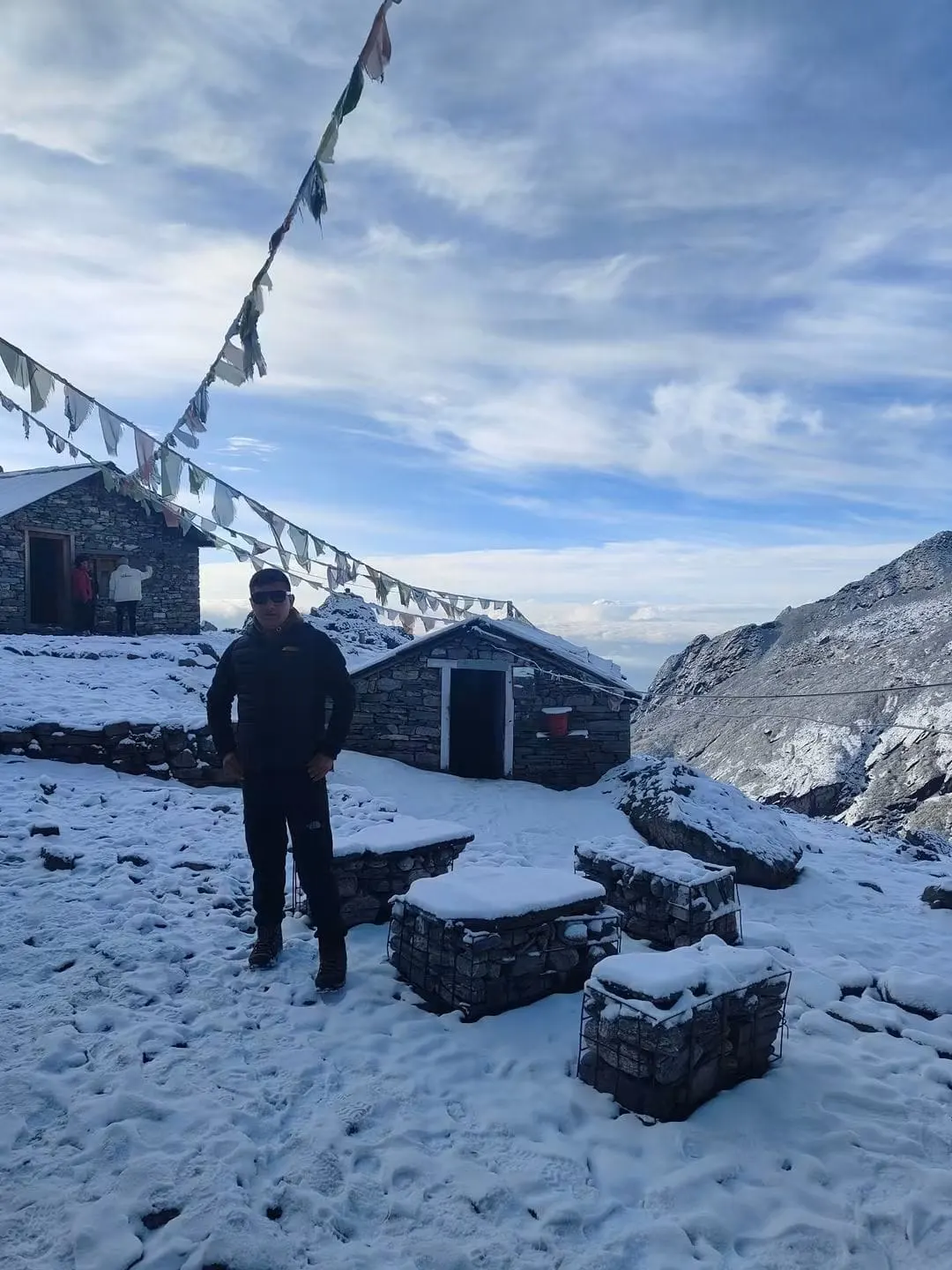 A man standing in the snow beside stone huts, with prayer flags overhead