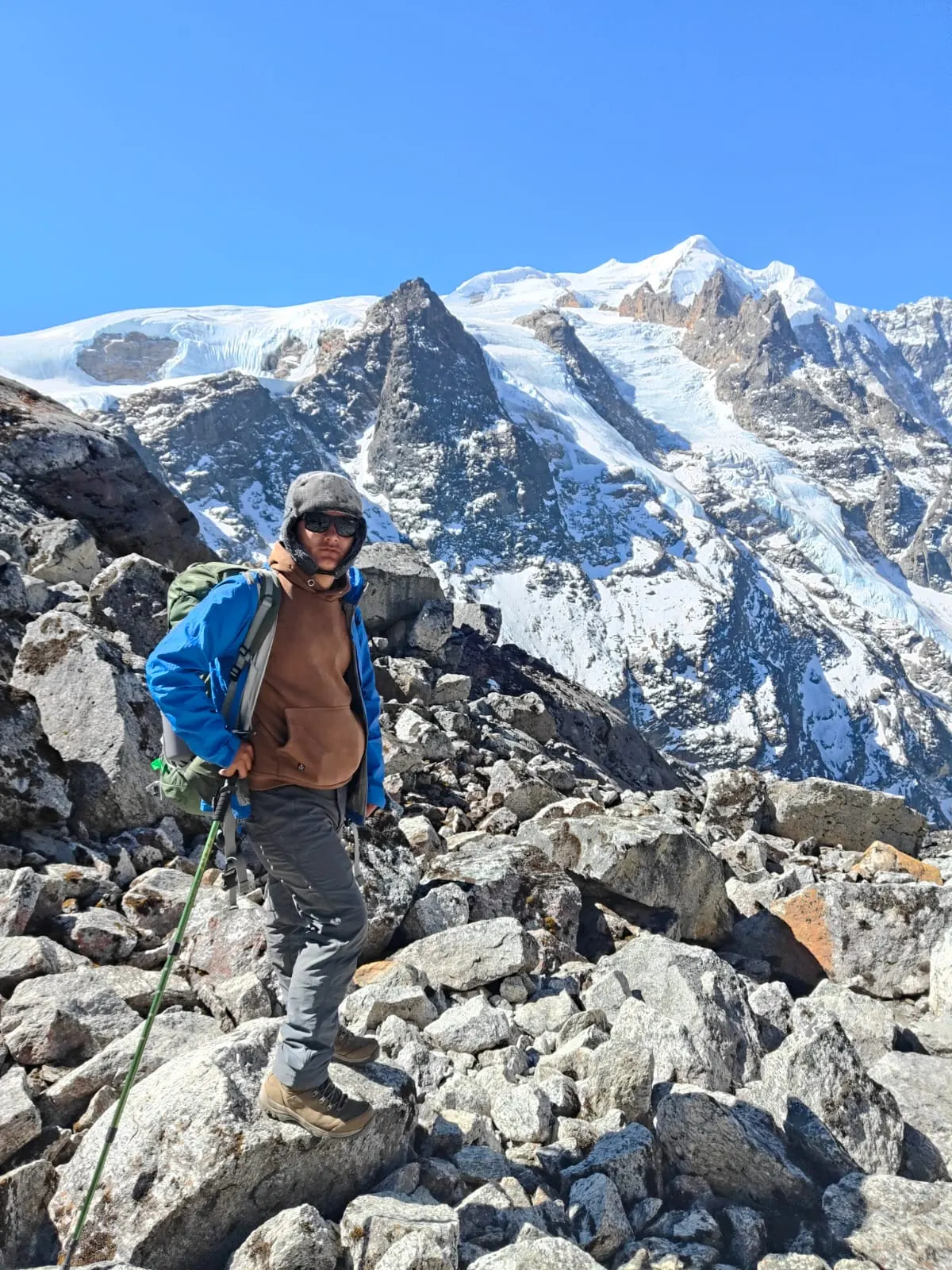 A trekker with walking poles on a rocky slope below a snow-covered peak