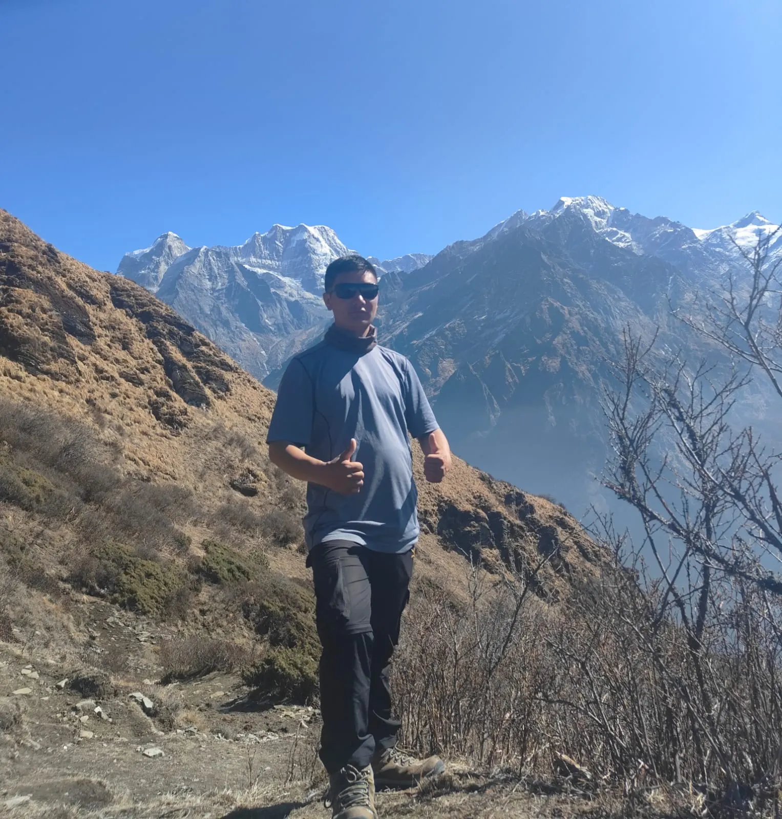 A man giving a thumbs-up on a dry hillside, with snow peaks behind