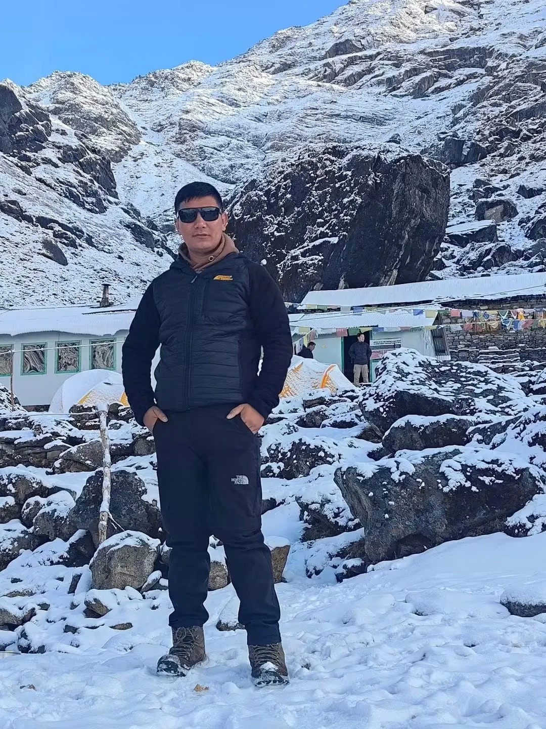 A man standing in the snow in front of lodges, with rocky peaks behind