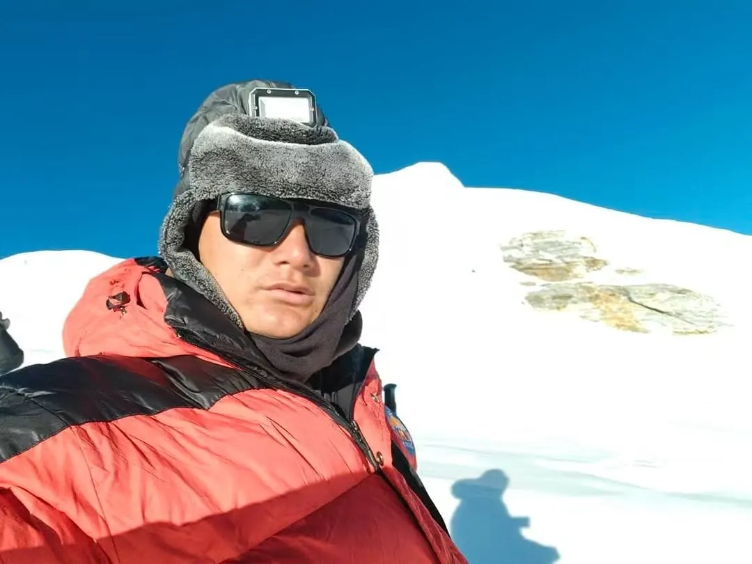 A close-up selfie of a climber in a red jacket, fur hat and dark glasses on a snowfield