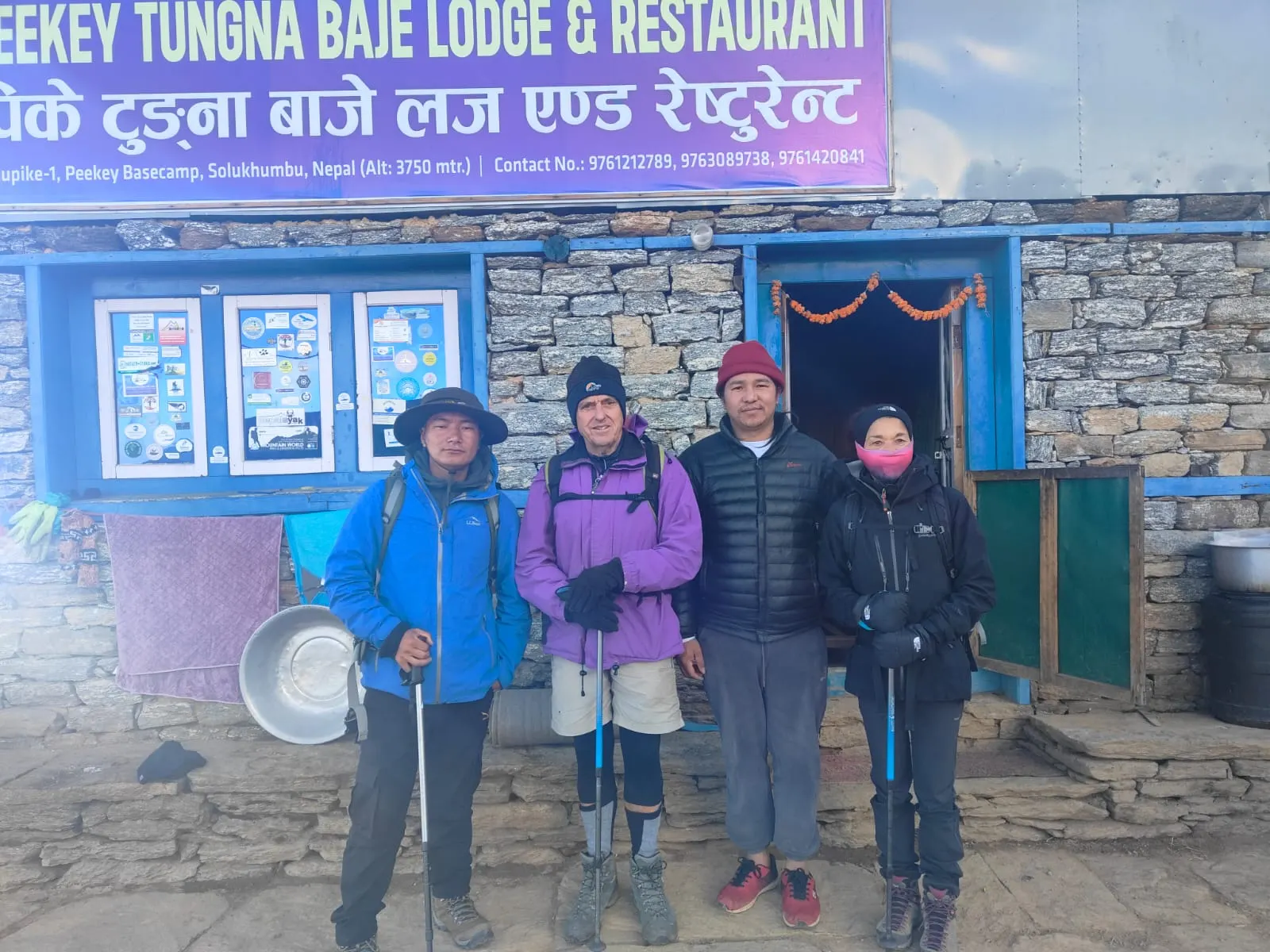 Four trekkers outside a stone lodge with a red signboard and blue windows