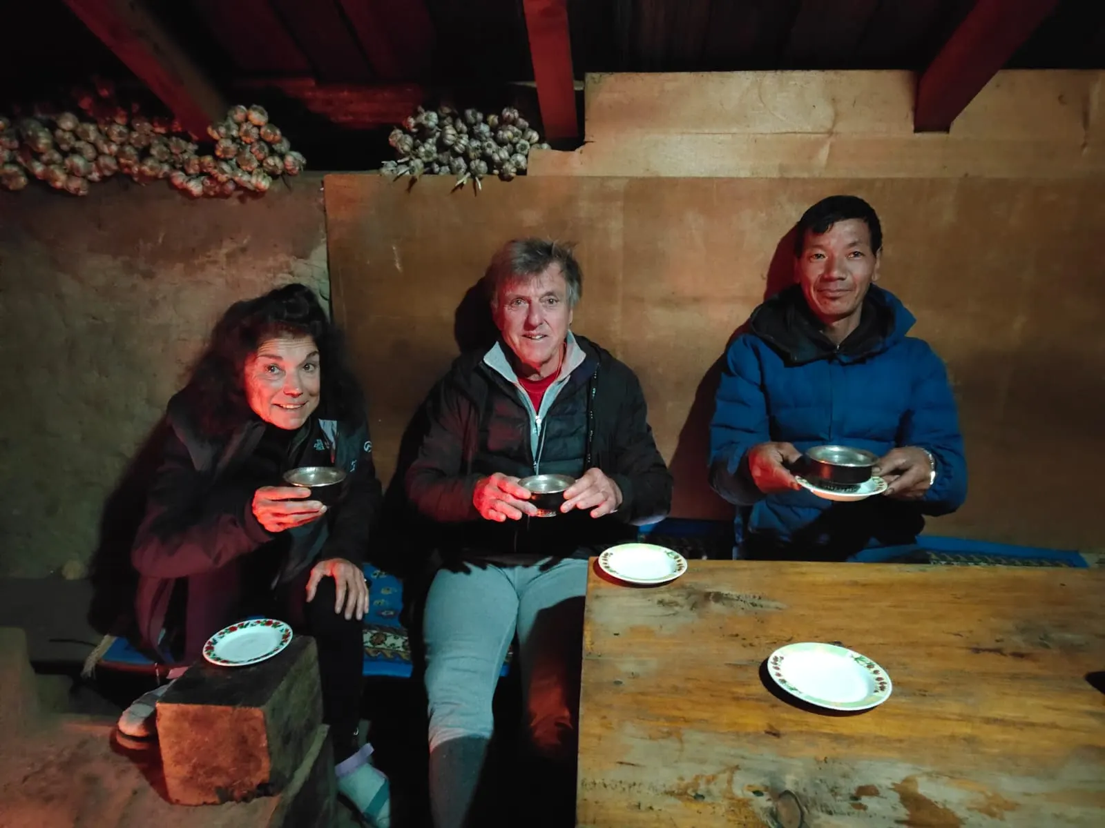 Three people sharing tea at a wooden table inside a lodge kitchen