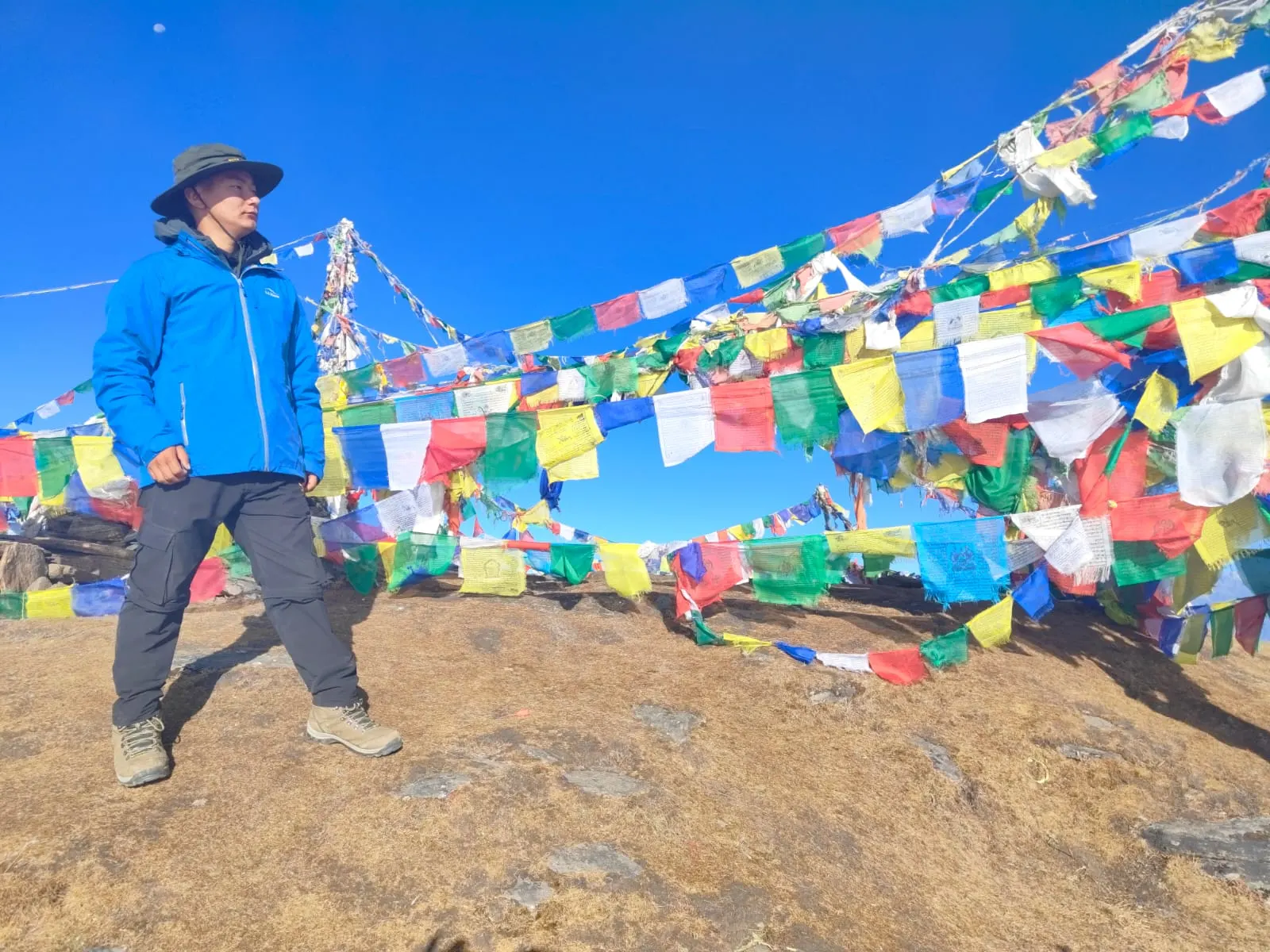 A man in a blue jacket standing among rows of colourful prayer flags on a hilltop